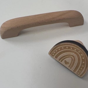 May include: Two wooden cabinet hardware pieces. One is a light brown, curved handle. The other is a half-circle wooden knob with a rainbow design, featuring hearts and arches. Both are on a white surface.