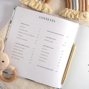 May include: A white baby memory book with a gold pen lying on top of it. The book's contents page is visible, listing sections like "Expecting You", "The First Weeks With You", and "Your First Years".