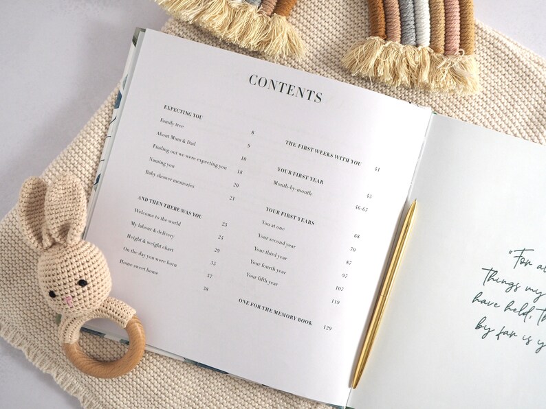 May include: A white baby memory book with a gold pen lying on top of it. The book's contents page is visible, listing sections like "Expecting You", "The First Weeks With You", and "Your First Years".