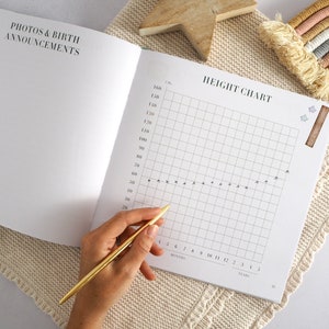 May include: A white baby book with a height chart on the right page. The chart has a grid with markings for months and years. A hand is holding a gold pen over the chart.