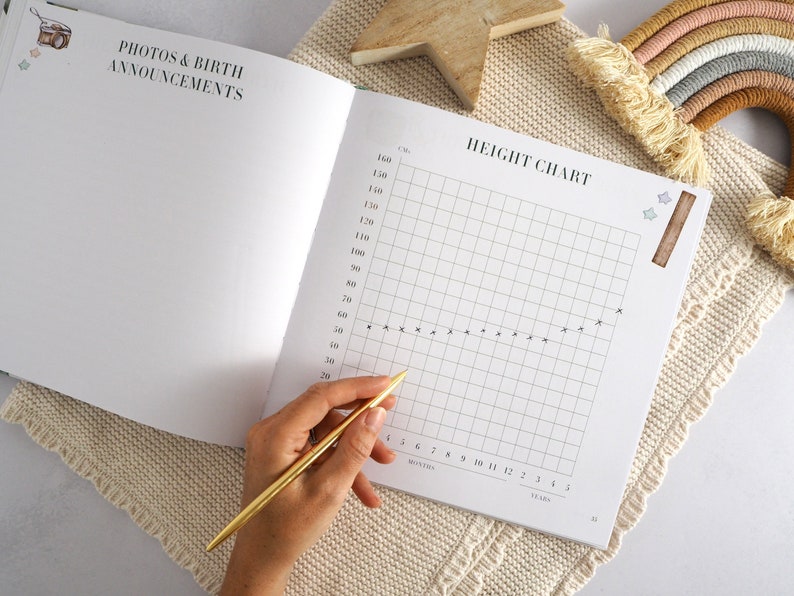 May include: A white baby book with a height chart on the right page. The chart has a grid with markings for months and years. A hand is holding a gold pen over the chart.