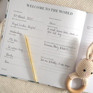 May include: A white baby book with a gold pen on top of it. The book has the words "Welcome to the World" on the cover. The book is open to a page with the baby's birth information, including their birth date, weight, length, and hair and eye color. The book also includes space for the names of the midwives or doctors who delivered the baby, as well as a section for the parents to write about their experience.