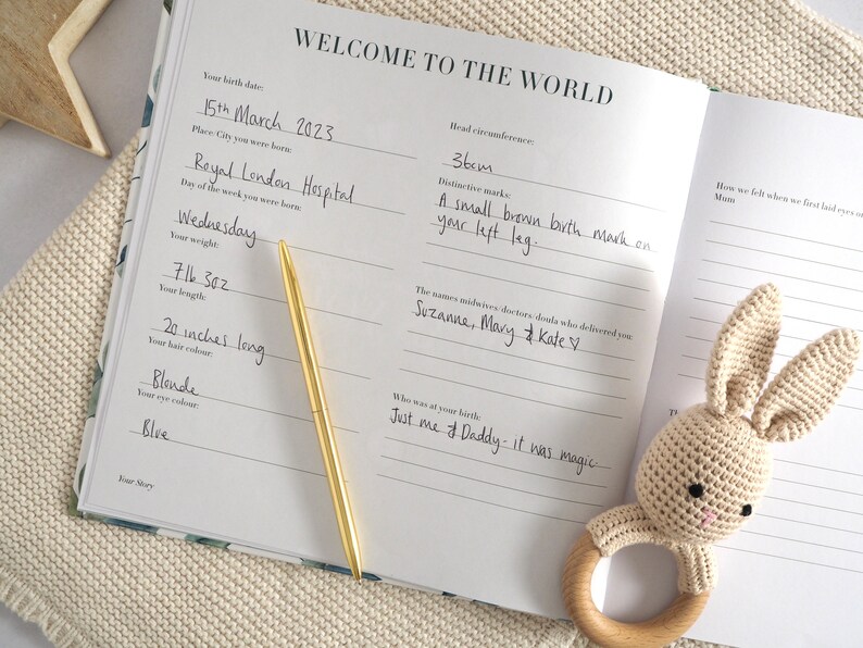 May include: A white baby book with a gold pen on top of it. The book has the words "Welcome to the World" on the cover. The book is open to a page with the baby's birth information, including their birth date, weight, length, and hair and eye color. The book also includes space for the names of the midwives or doctors who delivered the baby, as well as a section for the parents to write about their experience.
