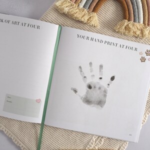 May include: A white book with the text "A work of art at four" and "Your hand print at four" on the cover. The book has a green spine and a handprint on the right page. There are small star and paw print stickers on the page.