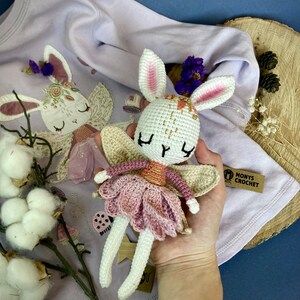 May include: A white crocheted bunny fairy doll wearing a pink tutu and gold glitter wings. The doll is holding a small flower in its hand. The doll is sitting on a purple shirt with a white crocheted bunny fairy design. The shirt has the text "Monys Crochet" on the bottom right corner.