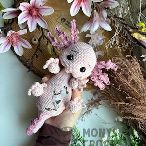 May include: A handmade pink crochet axolotl with black safety eyes and pink gills. The axolotl has small embroidered blue flowers on its side. The background includes a wooden slice, pink and white flowers, and greenery.