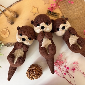 Pode incluir: Quatro pel&uacute;cias de lontra de croch&ecirc; feitas &agrave; m&atilde;o em cores marrom e creme. As lontras est&atilde;o deitadas de costas com as patas entrela&ccedil;adas. A imagem inclui um fundo de madeira, elementos decorativos e o texto "Monys Crochet".
