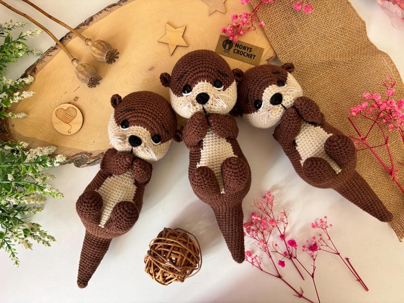 Pode incluir: Quatro pel&uacute;cias de lontra de croch&ecirc; feitas &agrave; m&atilde;o em cores marrom e creme. As lontras est&atilde;o deitadas de costas com as patas entrela&ccedil;adas. A imagem inclui um fundo de madeira, elementos decorativos e o texto "Monys Crochet".