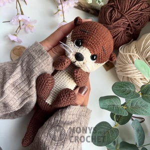 May include: A crocheted otter toy in shades of brown and cream, with black eyes and a black nose. The otter is being held. The image also shows brown and beige yarn. The text "MONYS CROCHET" is visible.