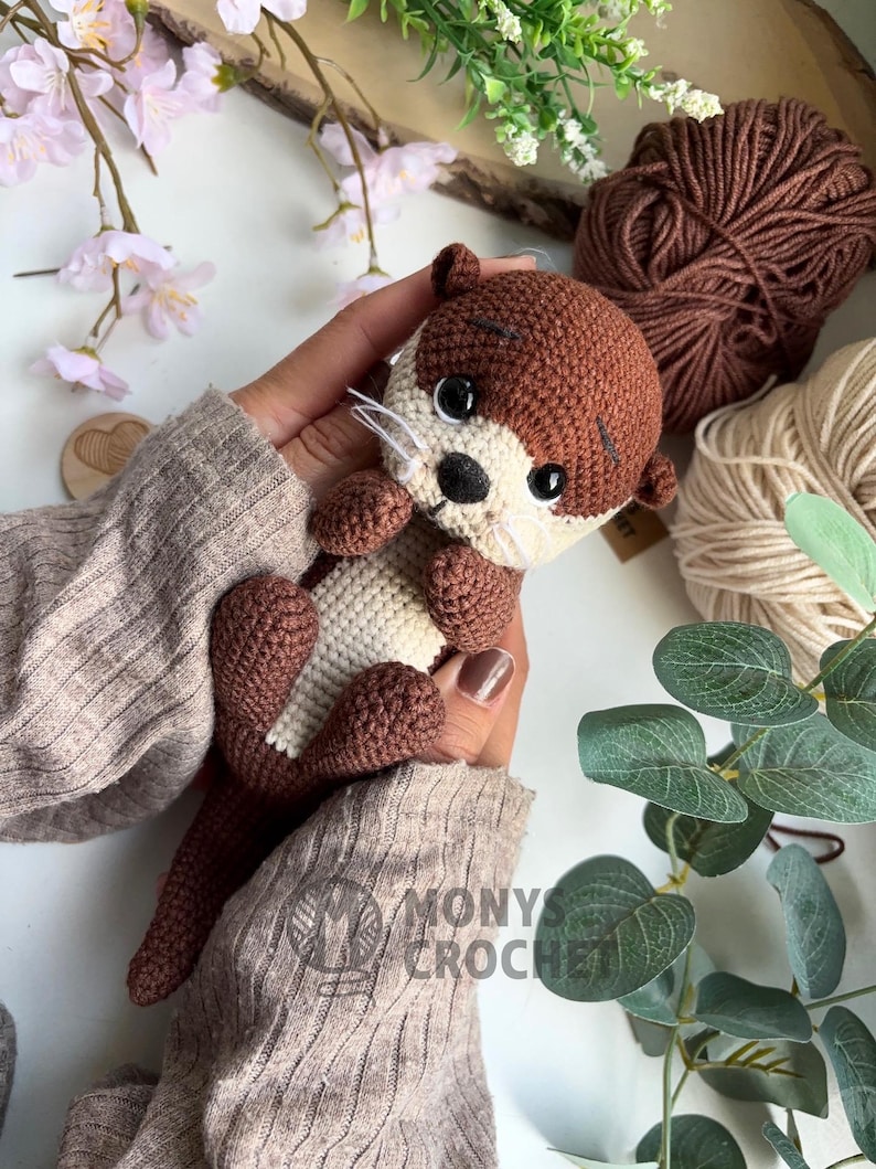 Pode incluir: Um bicho de pel&uacute;cia de lontra de croch&ecirc; feito &agrave; m&atilde;o, nas cores marrom e creme. A lontra tem olhos pretos, nariz preto e bigodes brancos. A imagem tamb&eacute;m mostra fios marrons e bege. O texto "MONYS CROCHET" &eacute; vis&iacute;vel.