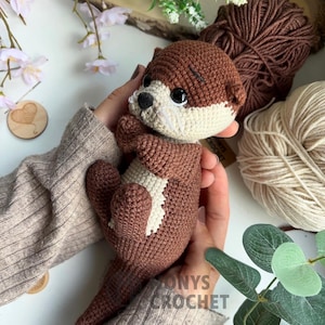 Pode incluir: Uma lontra de croch&ecirc; feita &agrave; m&atilde;o em tons de castanho e creme. O brinquedo de pel&uacute;cia apresenta um design detalhado com uma marca branca no peito e olhos pretos. A lontra &eacute; segurada em m&atilde;os, com fios e vegeta&ccedil;&atilde;o ao fundo.
