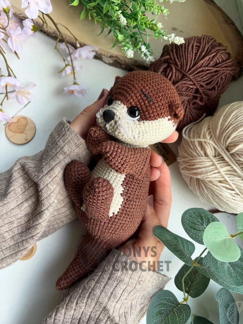 Pode incluir: Uma lontra de croch&ecirc; feita &agrave; m&atilde;o em tons de castanho e creme. O brinquedo de pel&uacute;cia apresenta um design detalhado com uma marca branca no peito e olhos pretos. A lontra &eacute; segurada em m&atilde;os, com fios e vegeta&ccedil;&atilde;o ao fundo.