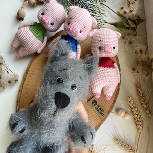 Puede incluir: Tres peluches de cerdo de crochet rosa con camisas de diferentes colores, una camisa verde, azul y roja. Una peluche de lobo de crochet gris con ojos negros y una nariz negra. Las peluches están sentadas sobre una superficie de madera con una etiqueta de papel marrón que dice "Monys Crochet".