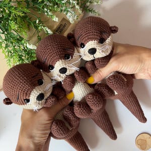 Pode incluir: S&atilde;o exibidas tr&ecirc;s lontras de croch&ecirc; feitas &agrave; m&atilde;o em tons de marrom e creme. As lontras t&ecirc;m olhos pretos, bigodes brancos e barriga creme. O texto "MONYS CROCHET" &eacute; vis&iacute;vel em um saco de papel marrom ao fundo.