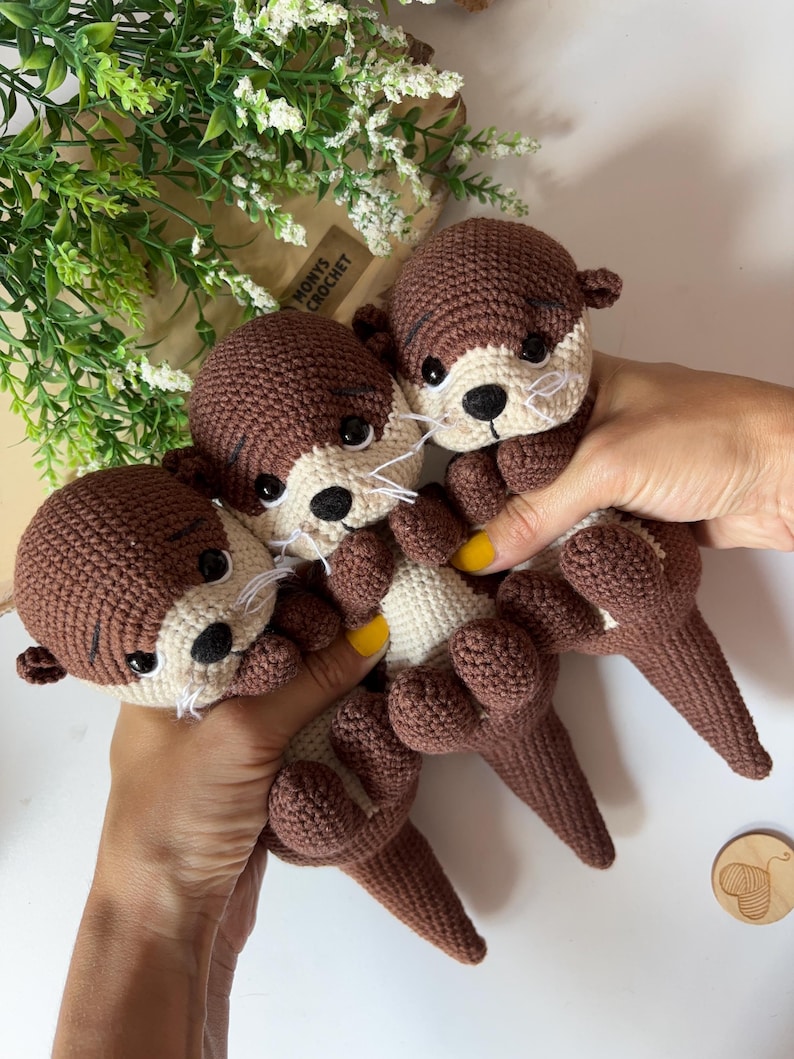 Pode incluir: S&atilde;o exibidas tr&ecirc;s lontras de croch&ecirc; feitas &agrave; m&atilde;o em tons de marrom e creme. As lontras t&ecirc;m olhos pretos, bigodes brancos e barriga creme. O texto "MONYS CROCHET" &eacute; vis&iacute;vel em um saco de papel marrom ao fundo.