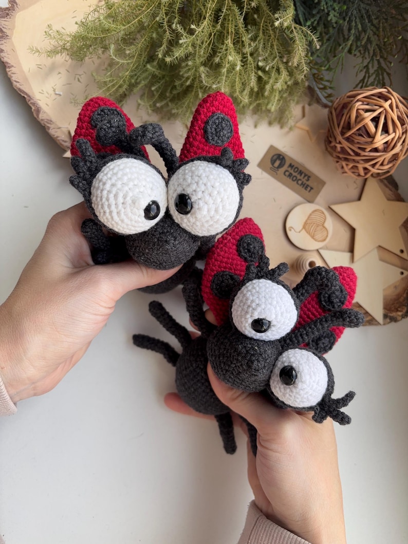 Puede incluir: Dos criaturas hechas a mano con ganchillo, con grandes ojos blancos, cuerpos negros y alas rojas y negras. Las criaturas se sostienen sobre un fondo neutro. El texto "MONTY'S CROCHET" es visible en una peque&ntilde;a etiqueta.