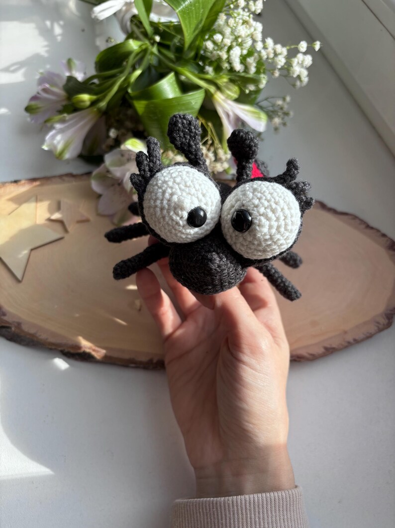 Puede incluir: Ara&ntilde;a negra de ganchillo hecha a mano con grandes ojos blancos y pupilas negras. La ara&ntilde;a tiene ocho patas y dos antenas. Un peque&ntilde;o detalle rojo es visible en la espalda de la ara&ntilde;a. Un art&iacute;culo decorativo caprichoso.