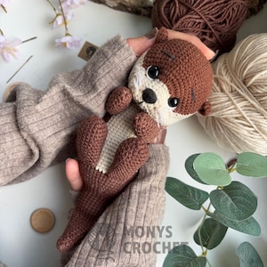 Pode incluir: Um brinquedo de lontra de croch&ecirc; feito &agrave; m&atilde;o em tons de marrom e creme, segurado em m&atilde;os. A lontra tem um rosto detalhado com olhos pretos e bigodes brancos. O texto "MONYS CROCHET" &eacute; vis&iacute;vel. Fios e vegeta&ccedil;&atilde;o no fundo.
