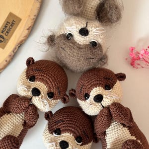 Pode incluir: Lontras de croch&ecirc; feitas &agrave; m&atilde;o em tons de castanho e creme. As lontras t&ecirc;m olhos pretos e bigodes brancos. A imagem mostra v&aacute;rias lontras em v&aacute;rias poses, mostrando os detalhes do design artesanal. O logotipo "MONYS CROCHET" &eacute; vis&iacute;vel.