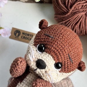 Pode incluir: Um brinquedo de pel&uacute;cia de lontra de croch&ecirc; feito &agrave; m&atilde;o, em tons de marrom e creme. A lontra tem olhos de seguran&ccedil;a pretos, um nariz preto e bigodes brancos. O brinquedo est&aacute; usando um su&eacute;ter cinza. Uma bola de l&atilde; marrom est&aacute; no fundo.
