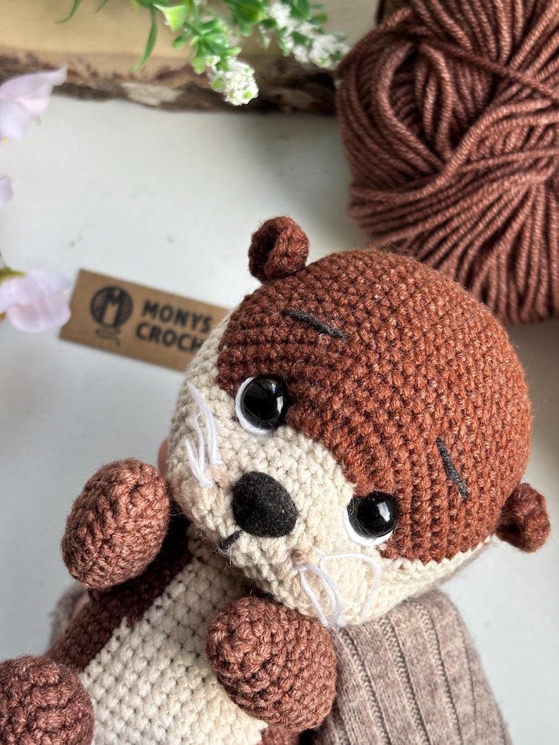 Pode incluir: Um brinquedo de pel&uacute;cia de lontra de croch&ecirc; feito &agrave; m&atilde;o, em tons de marrom e creme. A lontra tem olhos de seguran&ccedil;a pretos, um nariz preto e bigodes brancos. O brinquedo est&aacute; usando um su&eacute;ter cinza. Uma bola de l&atilde; marrom est&aacute; no fundo.