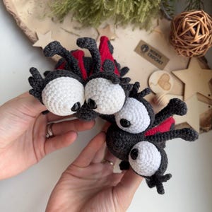 Puede incluir: Dos juguetes de mariquita hechos a ganchillo con grandes ojos blancos y pupilas negras, cuerpos gris oscuro y detalles de alas rojas. Las mariquitas tienen antenas finas y patas peque&ntilde;as, elaboradas con puntadas de hilo detalladas.