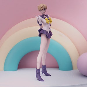 Puede incluir: Una figura de un personaje femenino de anime que lleva un atuendo de marinero blanco y morado con un lazo amarillo. Está de pie en una plataforma con un fondo de arcoíris.