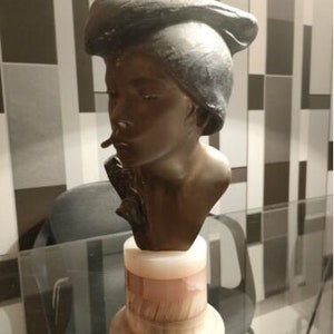 May include: A bronze sculpture of a woman's head wearing a hat, on a white marble base. The sculpture is on a glass table.