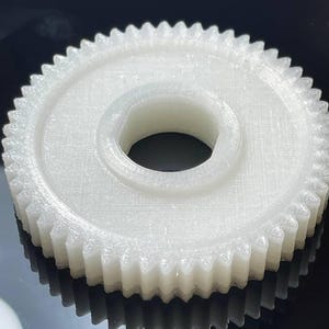 Puede incluir: Un engranaje de plástico blanco con un gran agujero central. El engranaje tiene una superficie lisa, ligeramente texturizada y una fila de dientes alrededor del borde exterior.