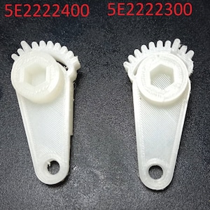 Puede incluir: Dos componentes de engranaje de plástico blanco con un centro hexagonal y un borde exterior dentado. Los componentes tienen una extensión en forma de asa con un agujero circular. El texto "5E2222400" y "5E2222300" está impreso encima.