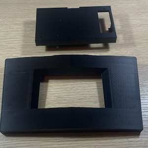 Peut inclure: Deux composants en plastique noir, l'un rectangulaire avec une découpe et l'autre plus petit, de forme rectangulaire. La pièce la plus grande a une ouverture rectangulaire au centre. Les deux pièces sont sur une surface brun clair.