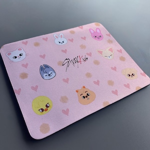 May include: A pink mouse mat with a repeating pattern of hearts and cartoon animal faces. The text "Stray Kids" is written in black in the centre of the mouse mat.