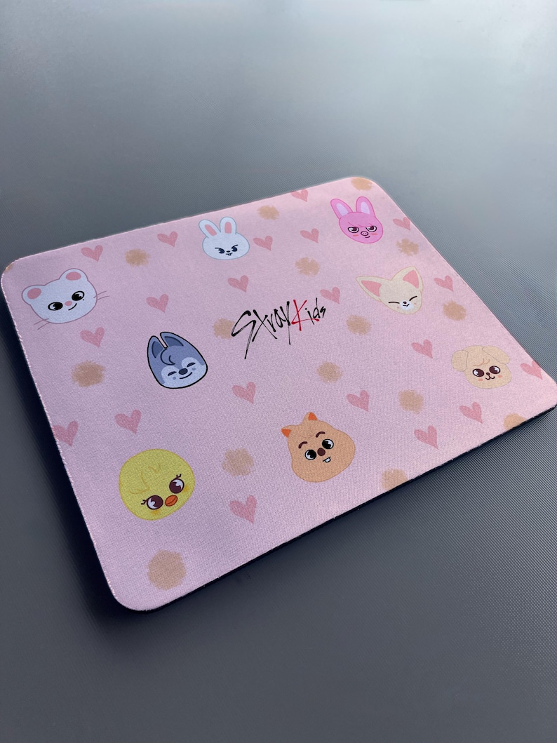 May include: A pink mouse mat with a repeating pattern of hearts and cartoon animal faces. The text "Stray Kids" is written in black in the centre of the mouse mat.