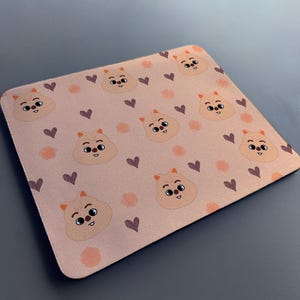 May include: A pink mouse mat with a repeating pattern of cartoon animals with brown eyes and hearts. The animals are light brown with orange accents.