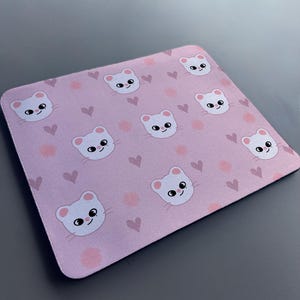 May include: A pink mouse mat with a repeating pattern of white cartoon cats and pink hearts.