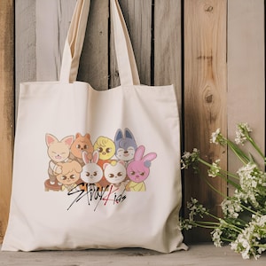 May include: A white canvas tote bag with a colorful cartoon image of eight animal characters. The characters are all wearing different outfits and have different expressions. The text "Stray Kids" is written in red and black below the image.
