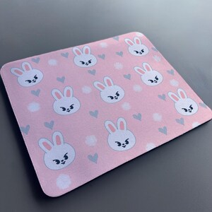 May include: A pink mouse mat with a repeating pattern of white bunnies with black eyes and grey ears. The bunnies are surrounded by small white dots and grey hearts.