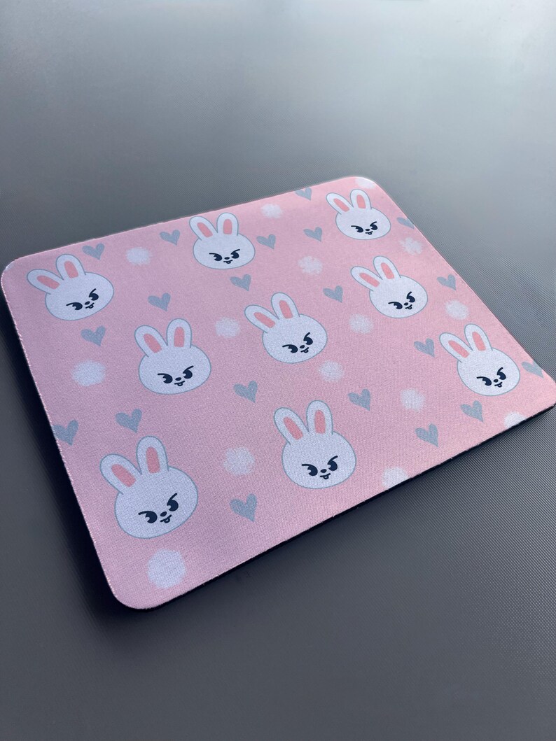 May include: A pink mouse mat with a repeating pattern of white bunnies with black eyes and grey ears. The bunnies are surrounded by small white dots and grey hearts.