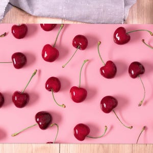 Pode incluir: Um fundo rosa com cerejas vermelhas espalhadas pela superfície. As cerejas estão dispostas em um padrão aleatório, com algumas sobrepostas.