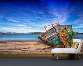 May include: A weathered wooden boat lies on a sandy beach with a blue sky and mountains in the background. A white chaise longue with a gold cushion sits in the foreground.