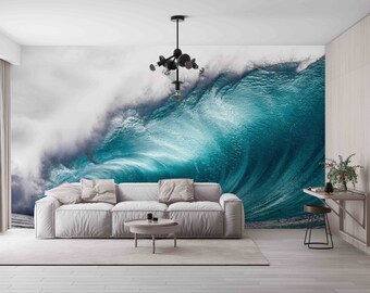 May include: A white couch with pillows sits in front of a wall mural depicting a large, crashing ocean wave. The wave is a vibrant blue and green color, with white foam at the crest. The couch is in a modern living room with a wooden floor and a simple black chandelier hanging from the ceiling.