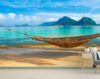 May include: A hammock hangs between two trees on a tropical beach with turquoise water and lush green mountains in the background.
