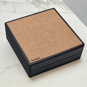 May include: A black square box with a cork top. The box has a two-part design with a lid and a base. The text "TrayLink" is visible on the front of the box. The cork top has a natural, textured appearance.