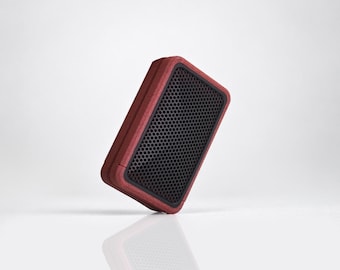 May include: A rectangular portable speaker with a deep red wooden frame and a black front panel featuring a fine honeycomb mesh grille. The device is positioned at an angle, showcasing its compact design and the contrast between the wood and metal.