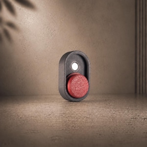 Op de afbeelding: Een donkergrijze, ovale deurklink met een rode, getextureerde knop. De klink heeft een witte, cirkelvormige accent in de buurt van de bovenkant. Het ontwerp is modern en minimalistisch, geschikt voor diverse interieurstijlen.