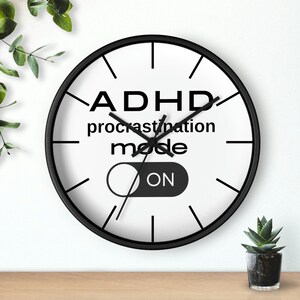 May include: A round wall clock with a black frame and a white face. The clock face features the text "ADHD procrastination mode ON" in black. Black lines mark the hours. A small succulent in a black pot is visible.