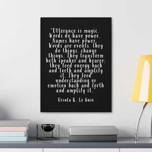 May include: A black canvas print with white text quoting Ursula K. Le Guin: "Utterance is magic. Words do have power..." The print is displayed on a shelf with books and a lamp.