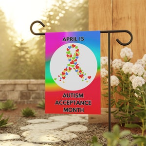 May include: A garden flag with a rainbow gradient background and the text "APRIL IS AUTISM ACCEPTANCE MONTH." A white circle features a ribbon made of colourful hearts. The flag is displayed on a black metal stand in a garden setting.