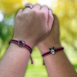 Pode incluir: Duas pulseiras de amizade com cord&atilde;o preto e rosa e um amuleto de um personagem de desenho animado. Os amuletos s&atilde;o Hello Kitty e Kuromi.