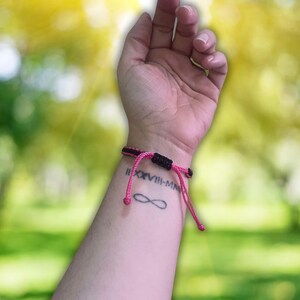 Pode incluir: Uma pulseira de corda rosa e preta com a inscri&ccedil;&atilde;o em algarismos romanos "II.XVIII-MM" e uma tatuagem de s&iacute;mbolo de infinito no pulso.